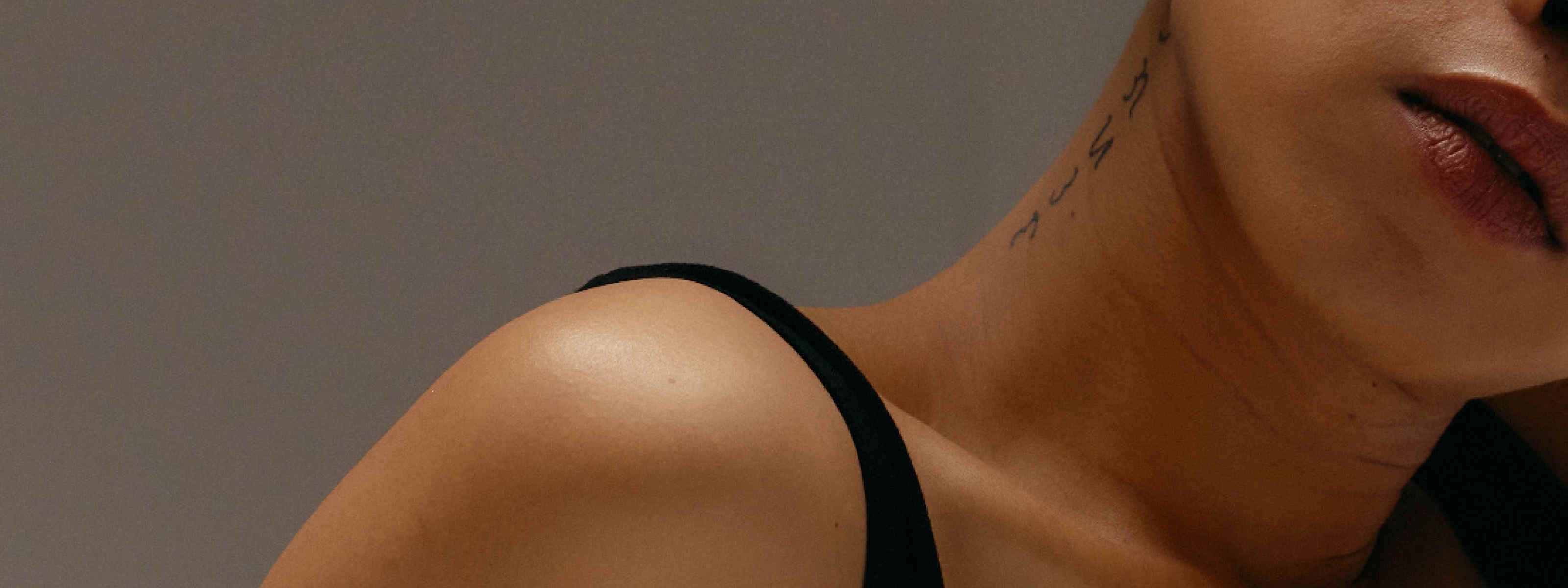 Close-up photo of a woman's shoulder and neck, wearing a black strap top, with a small script-style tattoo visible on the side of her throat.