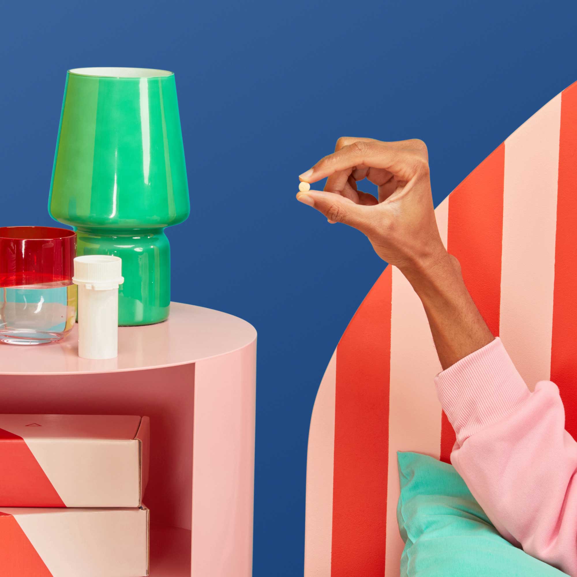 A man's hand holding a DoxyPEP pill near a pink nightstand with a pill vial, green lamp and a glass of water on top