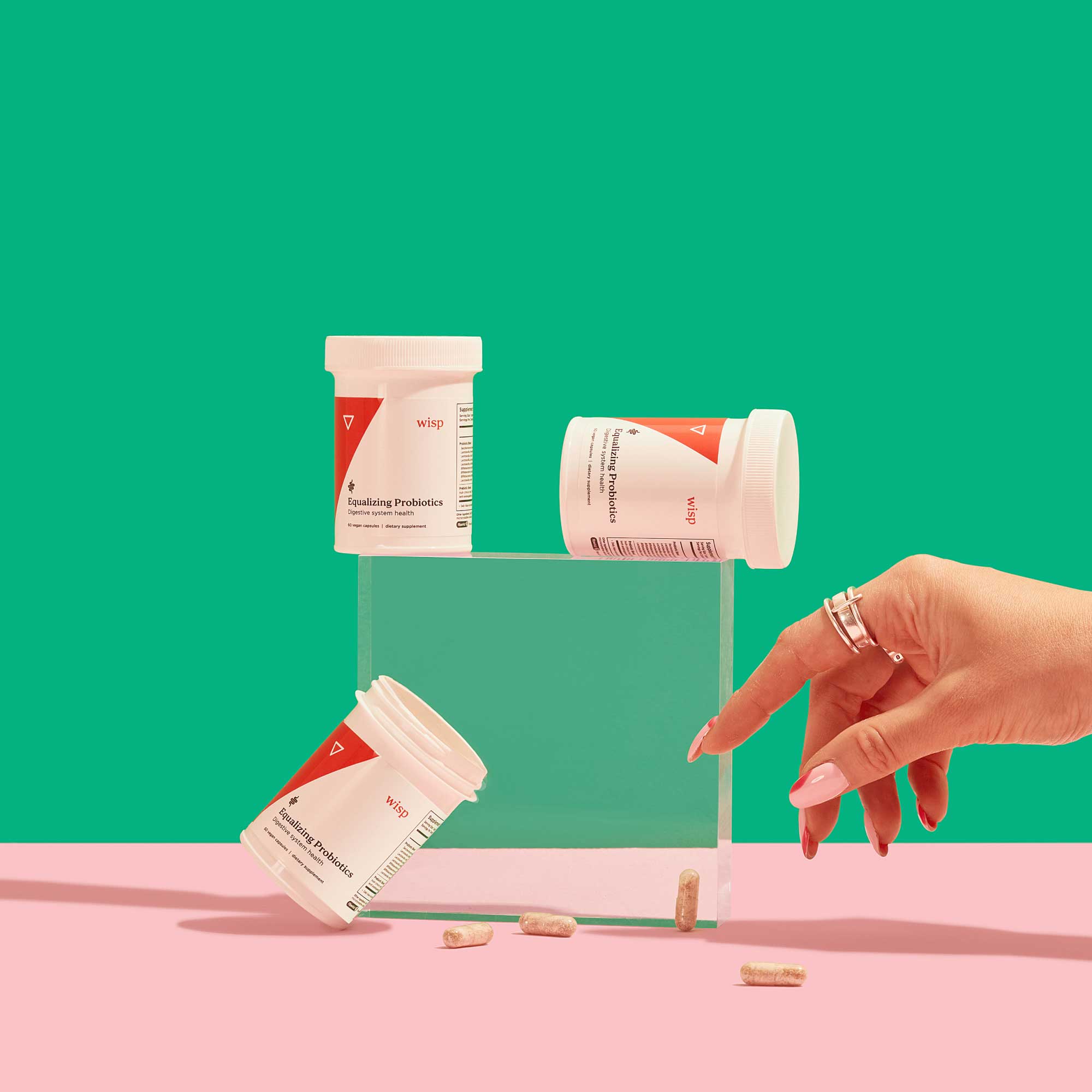 3 bottles of Wisp Equalizing Probiotics balanced on a clear square shape with a woman's hand reaching for spilled pills on a pink surface with a green background