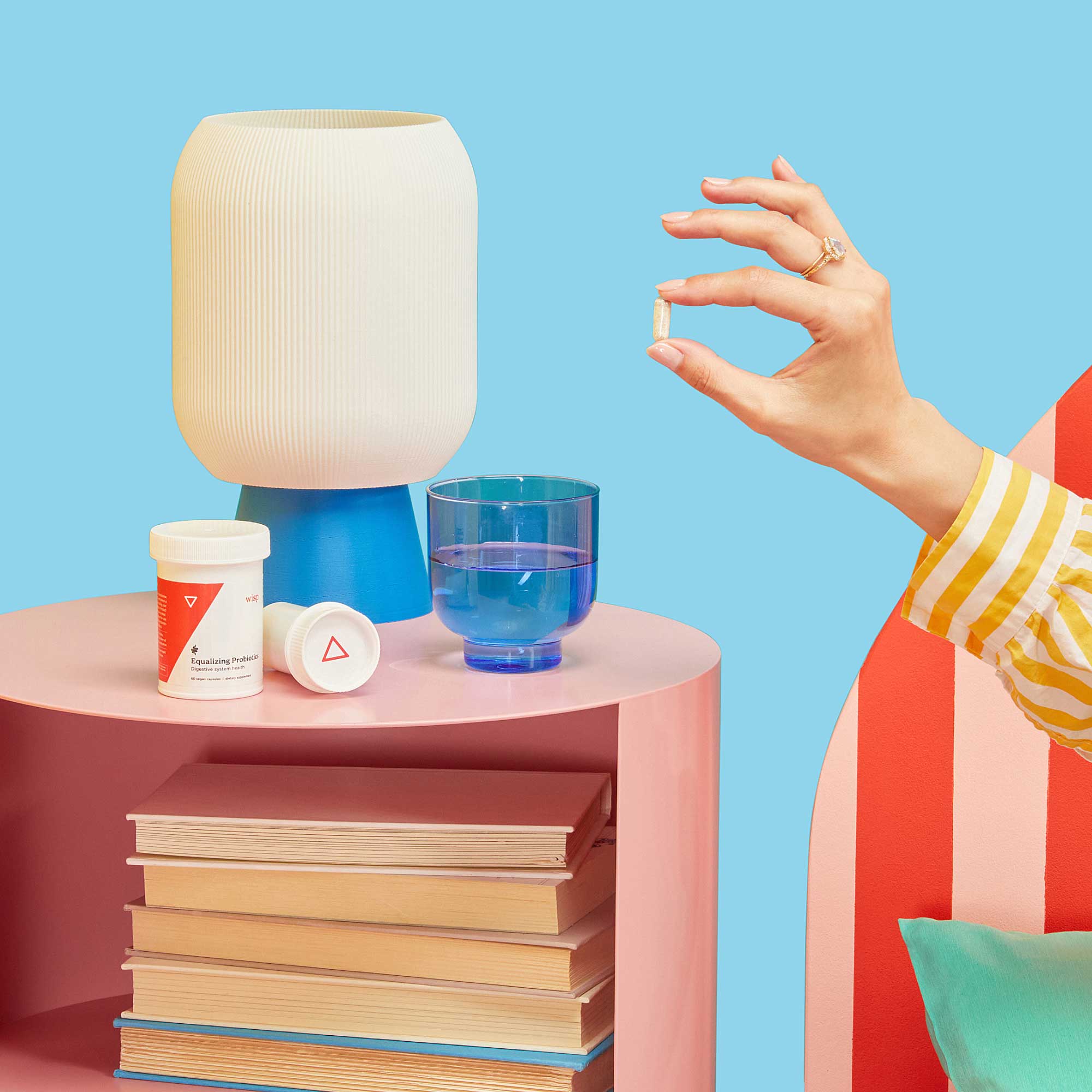 A woman's hand holding a probiotic pill near a pink nightstand with Wisp medication, a glass of water, and a lamp on top with books stacked inside in front of a light blue background