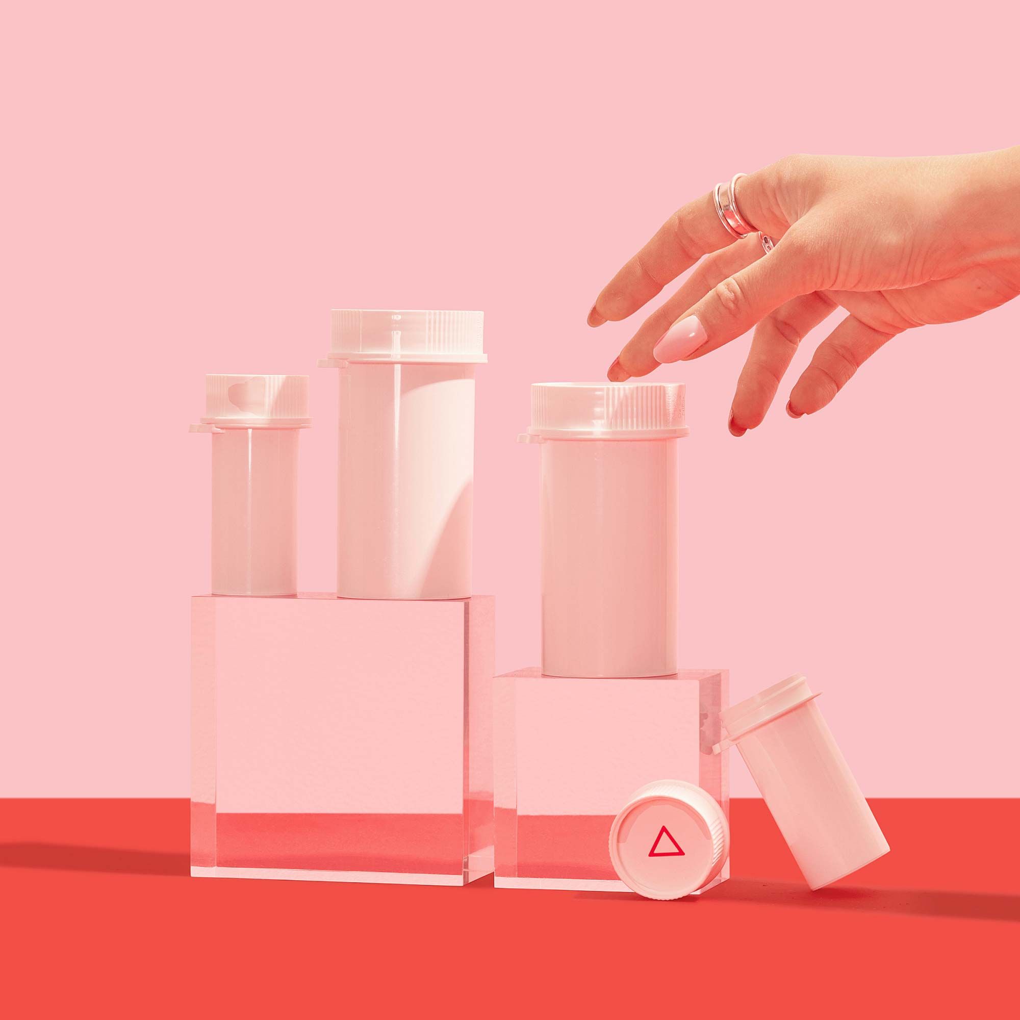 A woman's hand reaching for various Wisp medication bottles balanced on clear abstract shapes on a red surface with a pink background