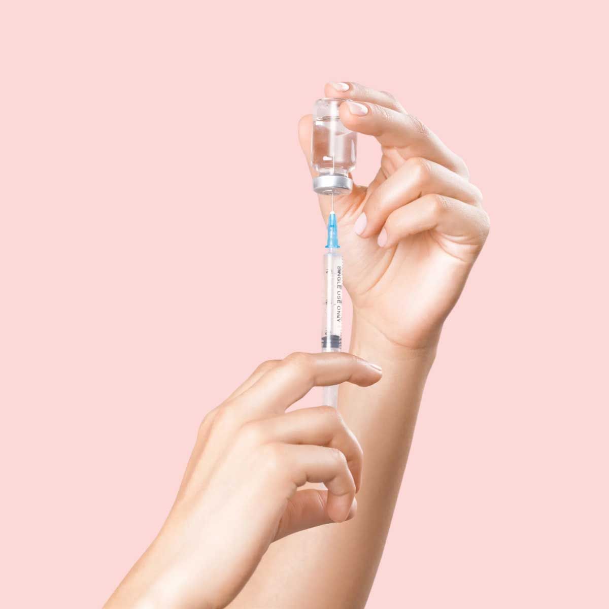 A person's hands filling a syringe with liquid from a vial in front of a pink background
