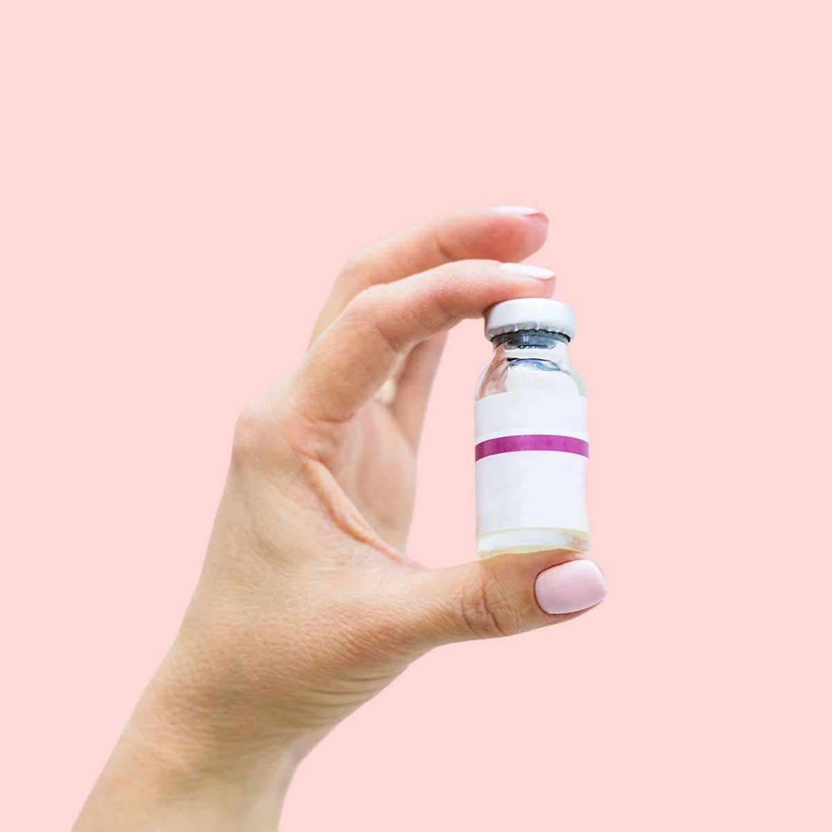 A person's hand holding an injection vial in front of a pink background