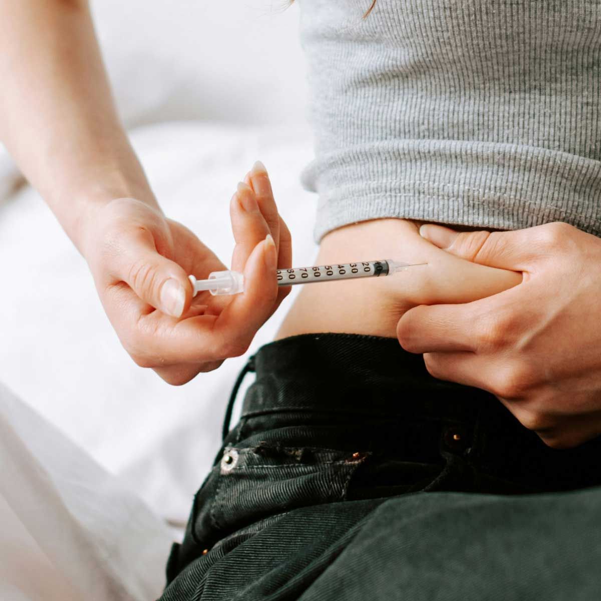 A person wearing a gray sweater and black jeans is self-administering an injection into their abdomen