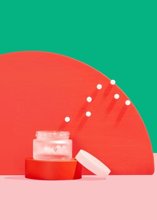 A small Wisp glass pill jar with pills floating out and colorful abstract shapes on a pink surface with a green background