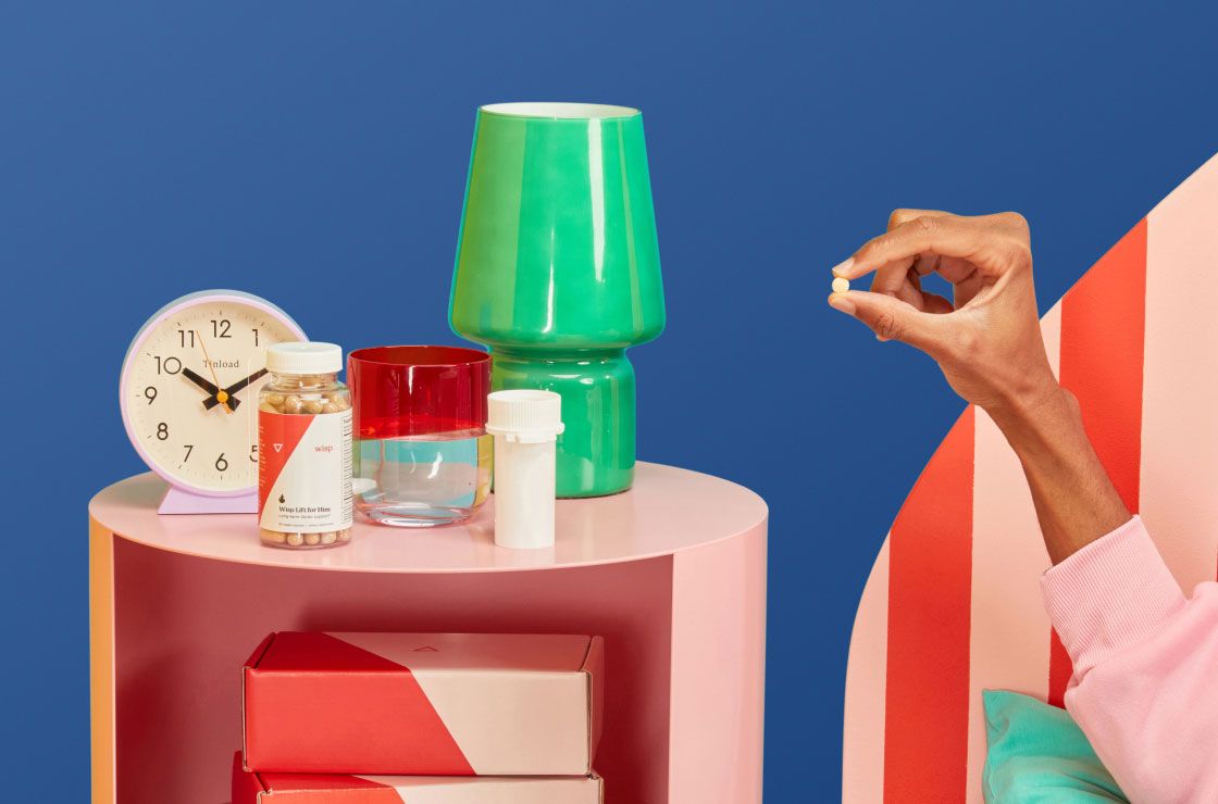 A man's hand holding a DoxyPEP pill next to a pink nightstand with a Wisp plastic pill Vial, Wisp Lift for Him, a green lamp, alarm clock, and a glass of water in front of a dark blue background