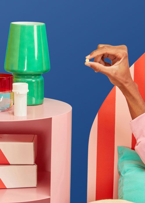 A man's hand holding a DoxyPEP pill near a pink nightstand with a Wisp pill vial, a green lamp and Wisp shipping boxes with a dark blue background