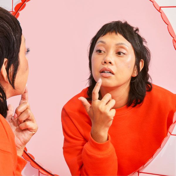 A woman wearing a red sweater is applying a hydrocolloid patch to her chin in a bathroom mirror with a pink background