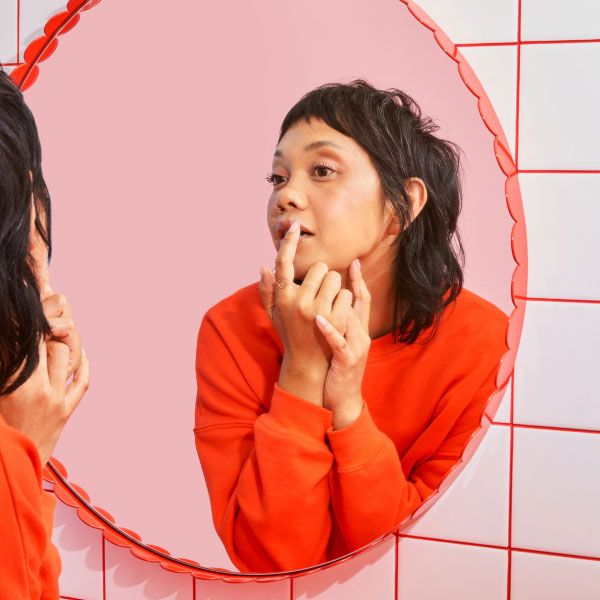 A woman wearing a red sweater is applying a hydrocolloid patch to her lip in a bathroom mirror with a pink background