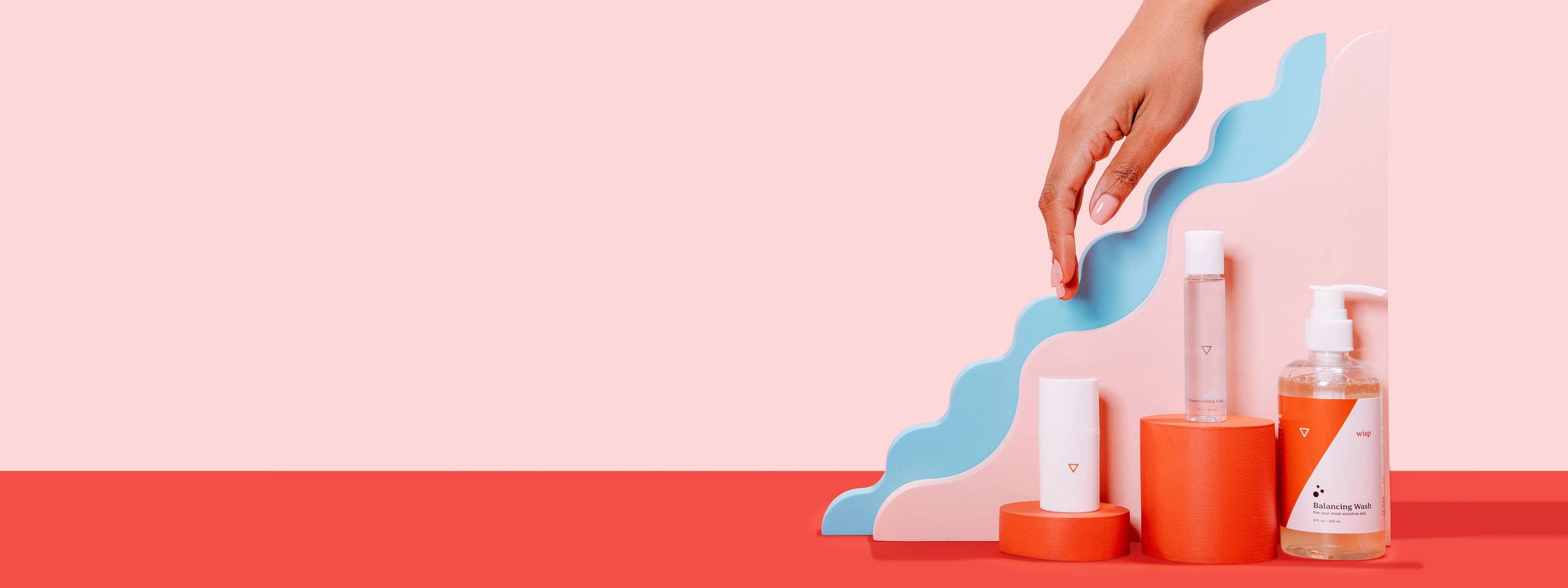 A woman's hand reaching for Wisp's OMG! Cream, Harmonizing Lube, and Balancing Wash balanced on colorful abstract shapes on a red surface with a pink background