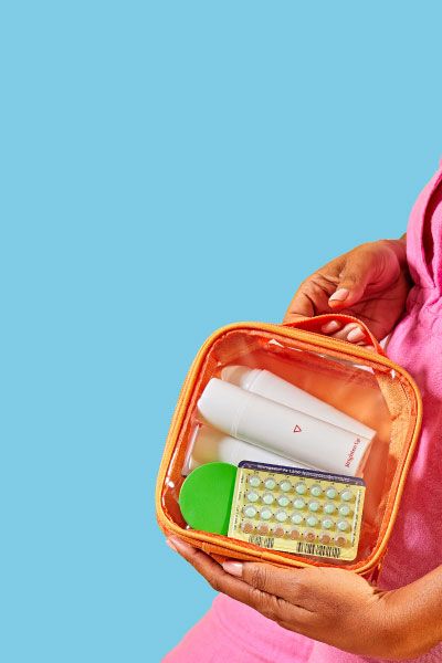 A woman wearing a pink jumpsuit and carrying a green travel bag is holding an orange pouch with Wisp birth control and skincare products in front of a light blue background