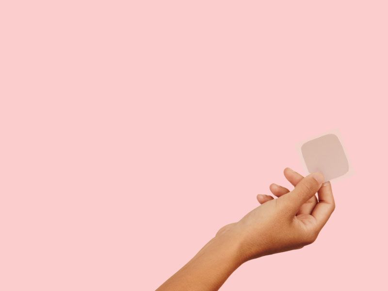 hand holding a birth control patch in front of a pink background