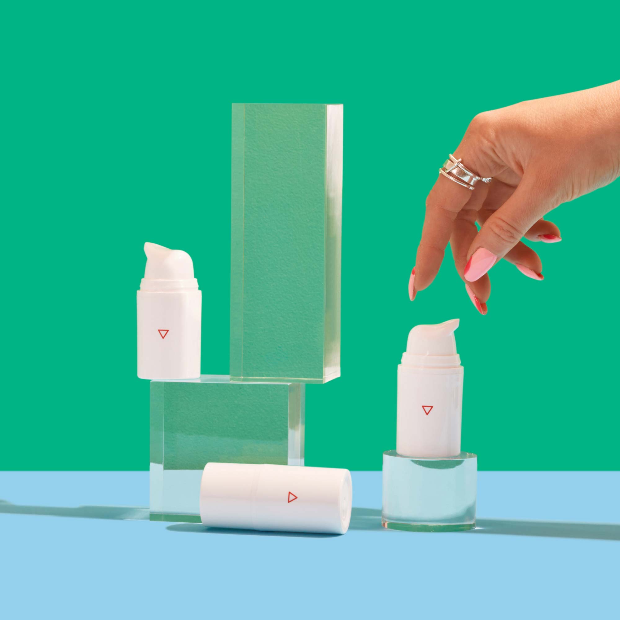 A woman's hand reaching for Wisp pump bottles balanced on clear abstract shapes on a light blue surface with a green background