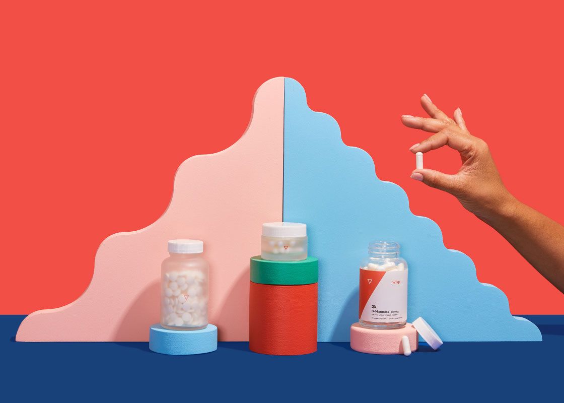 A woman's hand holding a pill next two various Wisp products balanced on colorful abstract shapes on a blue surface with a red background