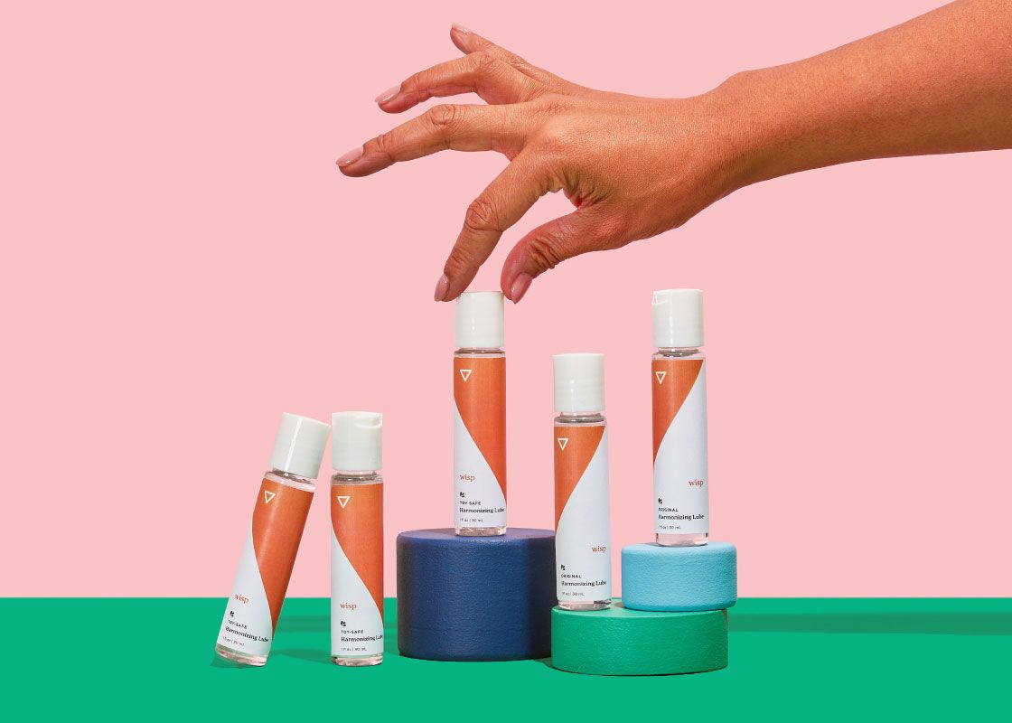 A woman's hand reaching for multiple Harmonizing Lube bottles balanced on colorful abstract shapes on a green surface with a pink background