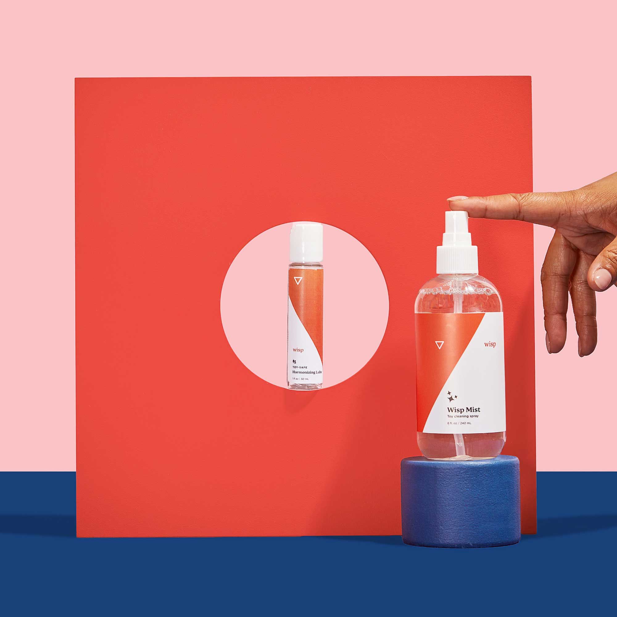 A woman's hand reaching for Wisp Mist balanced on a colorful abstract shape with Wisp Harmonizing Lube on a blue surface with a pink background