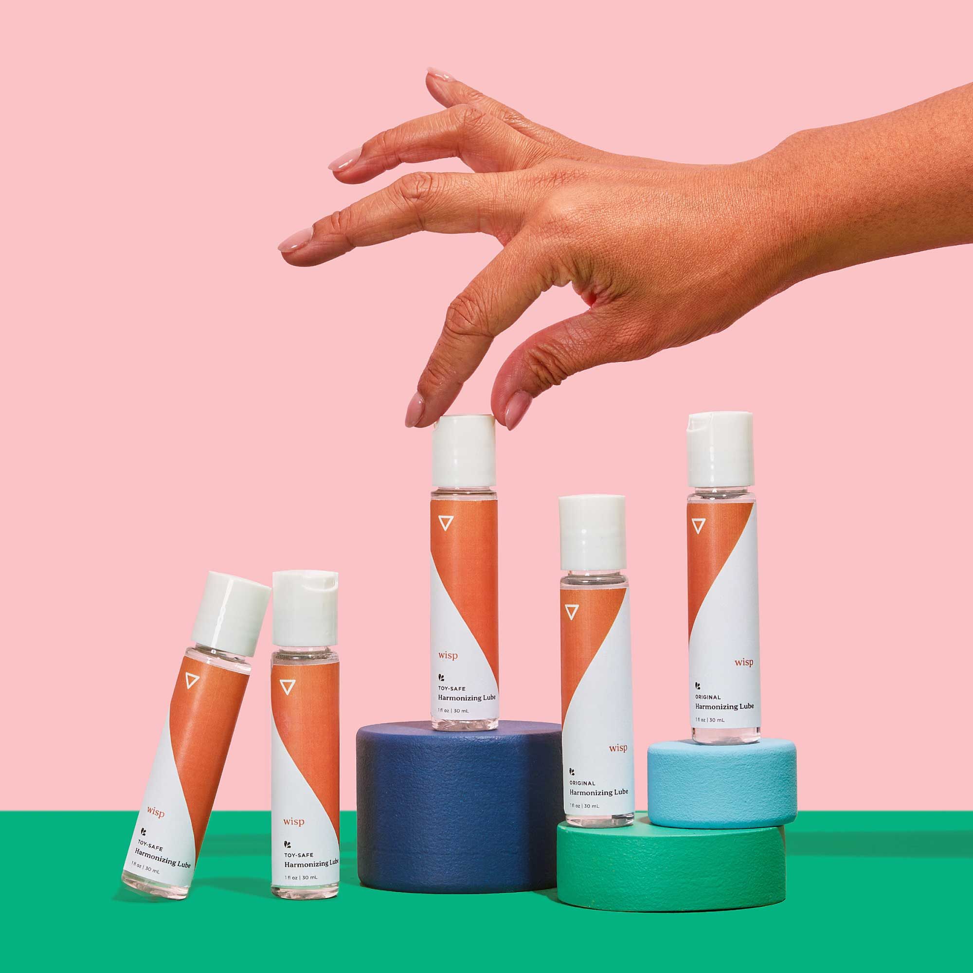 A woman's hand reaching for 5 Wisp Harmonizing Lubes balanced on colorful abstract shapes on a green surface with a pink background