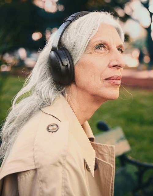 A woman with gray hair wearing headphones and a trench coat outdoors
