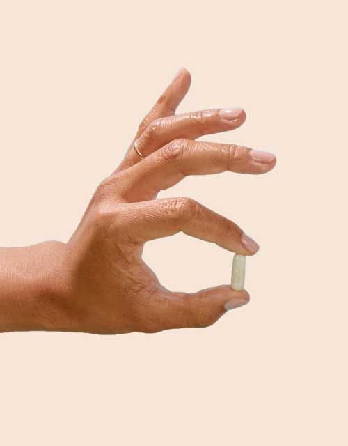 A person's hand holding a pill in front of a cream background