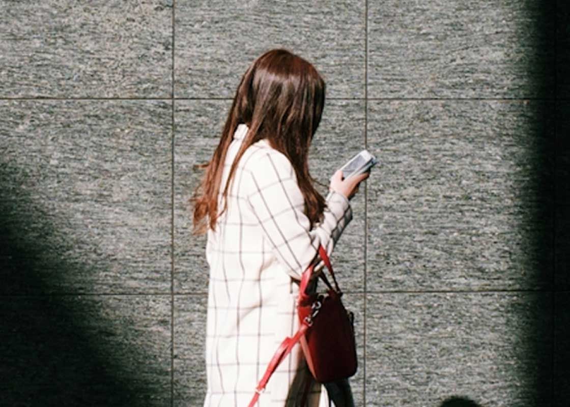 A woman with brown hair wearing a white dress is walking in a city while looking at her phone