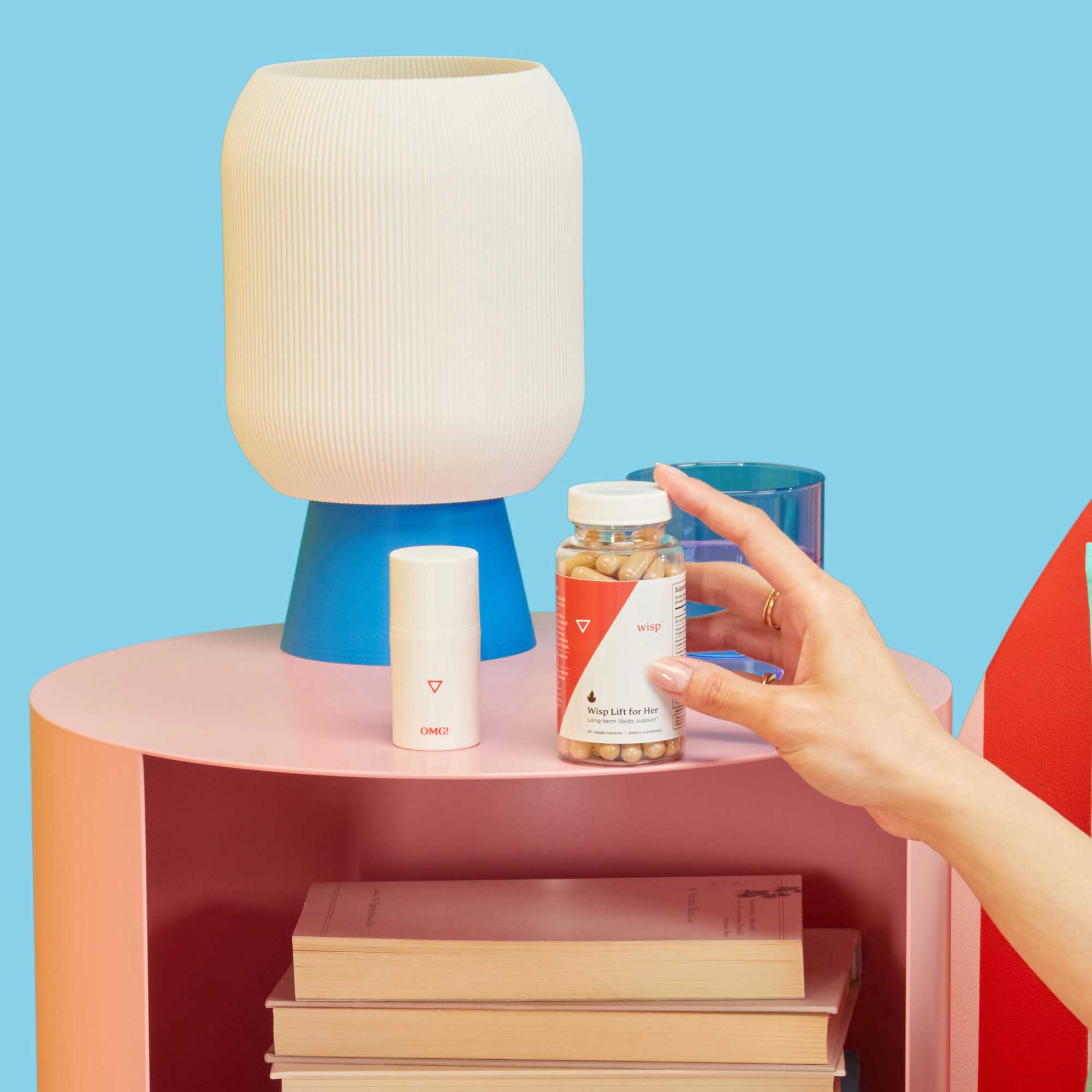A woman's hand reaching for a bottle of Wisp Lift for Her on a pink nightstand with a blue lamp and a glass of water in front of a light blue background