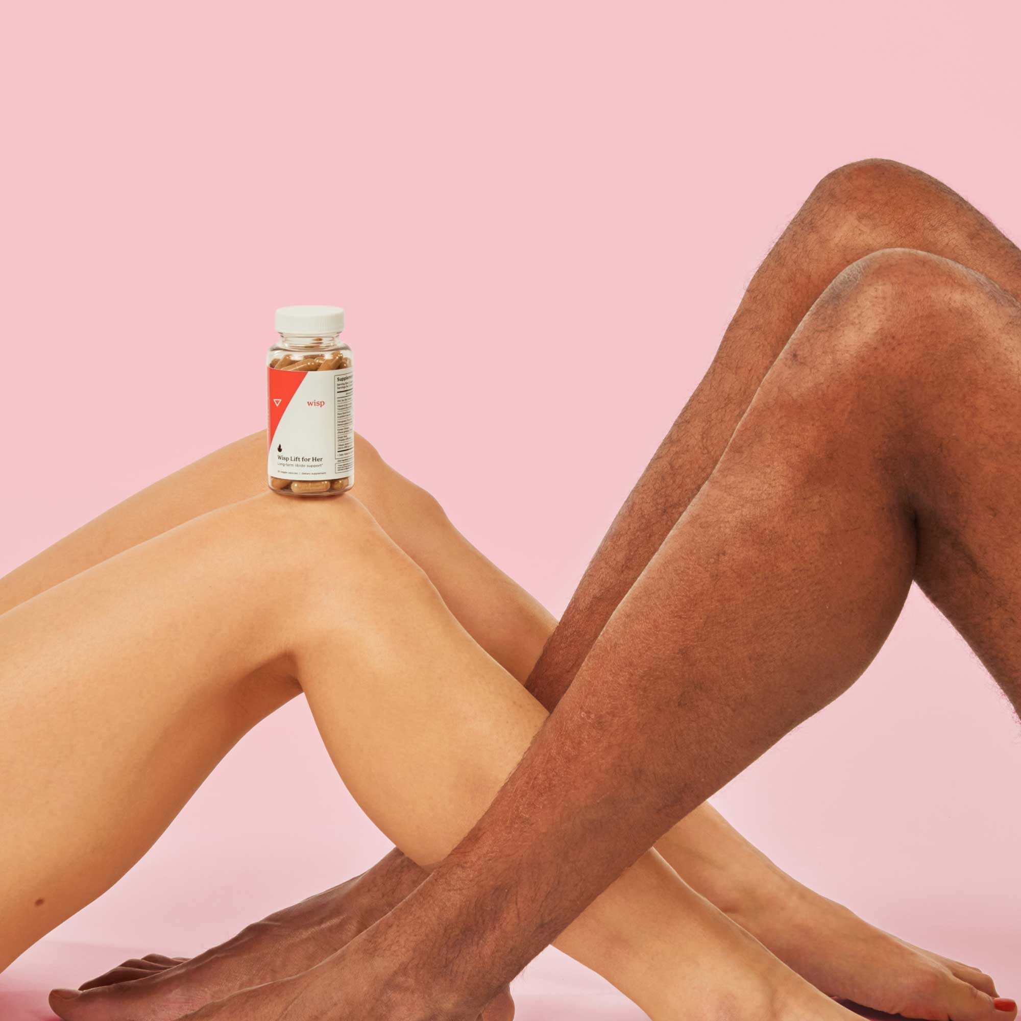 Intertwined legs with a bottle of Wisp Lift for Her balanced on on of their knees in front of a pink background