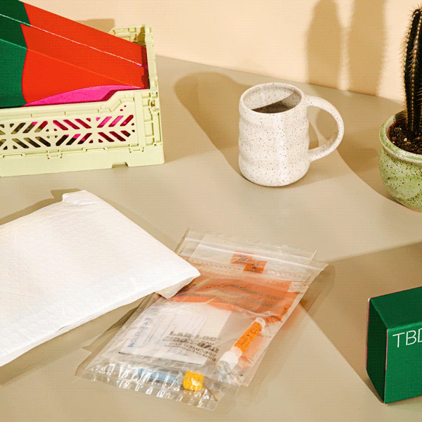 A person putting their testing samples into a mail envelope with TBD health shipping boxes, a coffee mug and a cactus on a tan surface