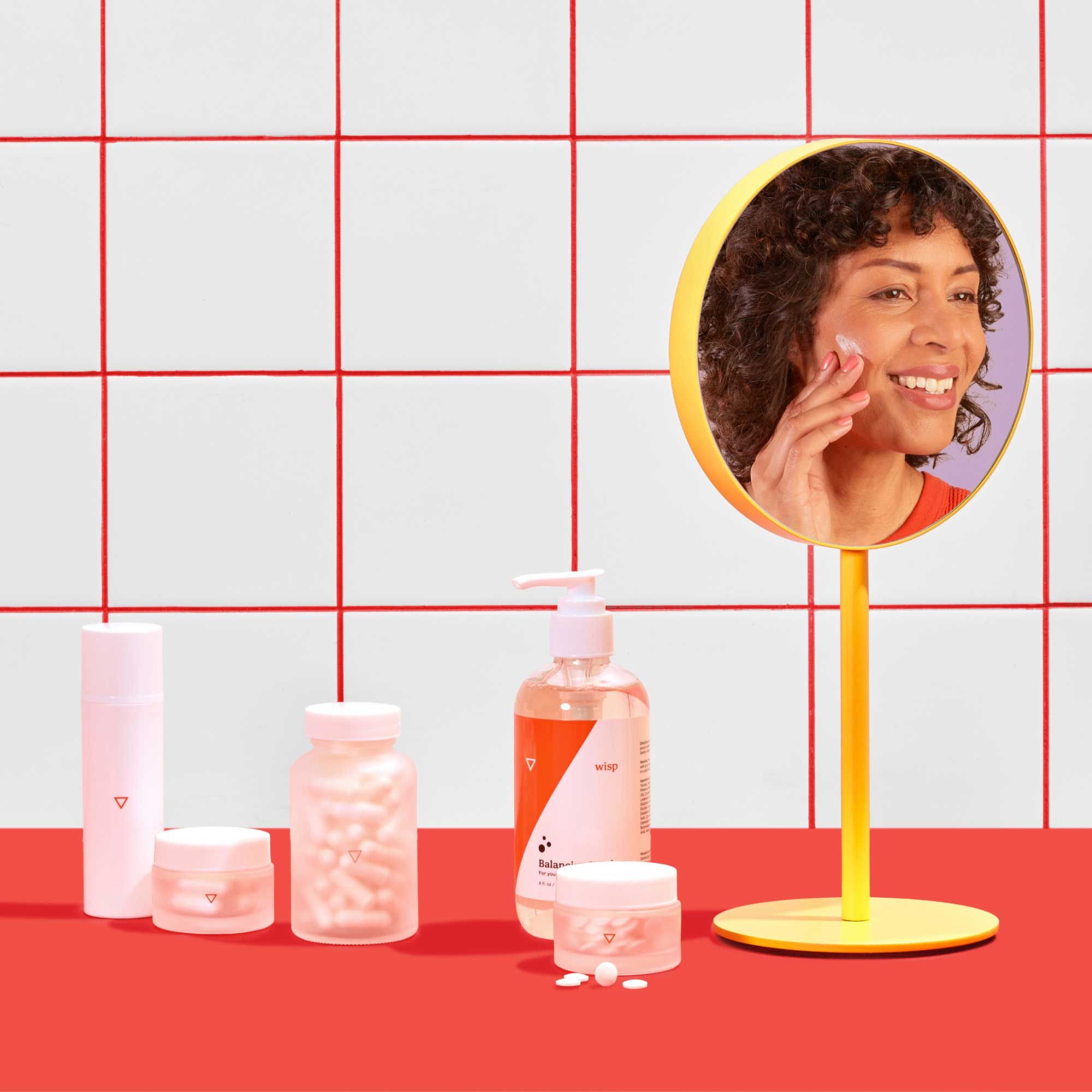 Various Wisp products on a red surface with a a woman's reflection in a yellow hand mirror applying Estriol Face Cream