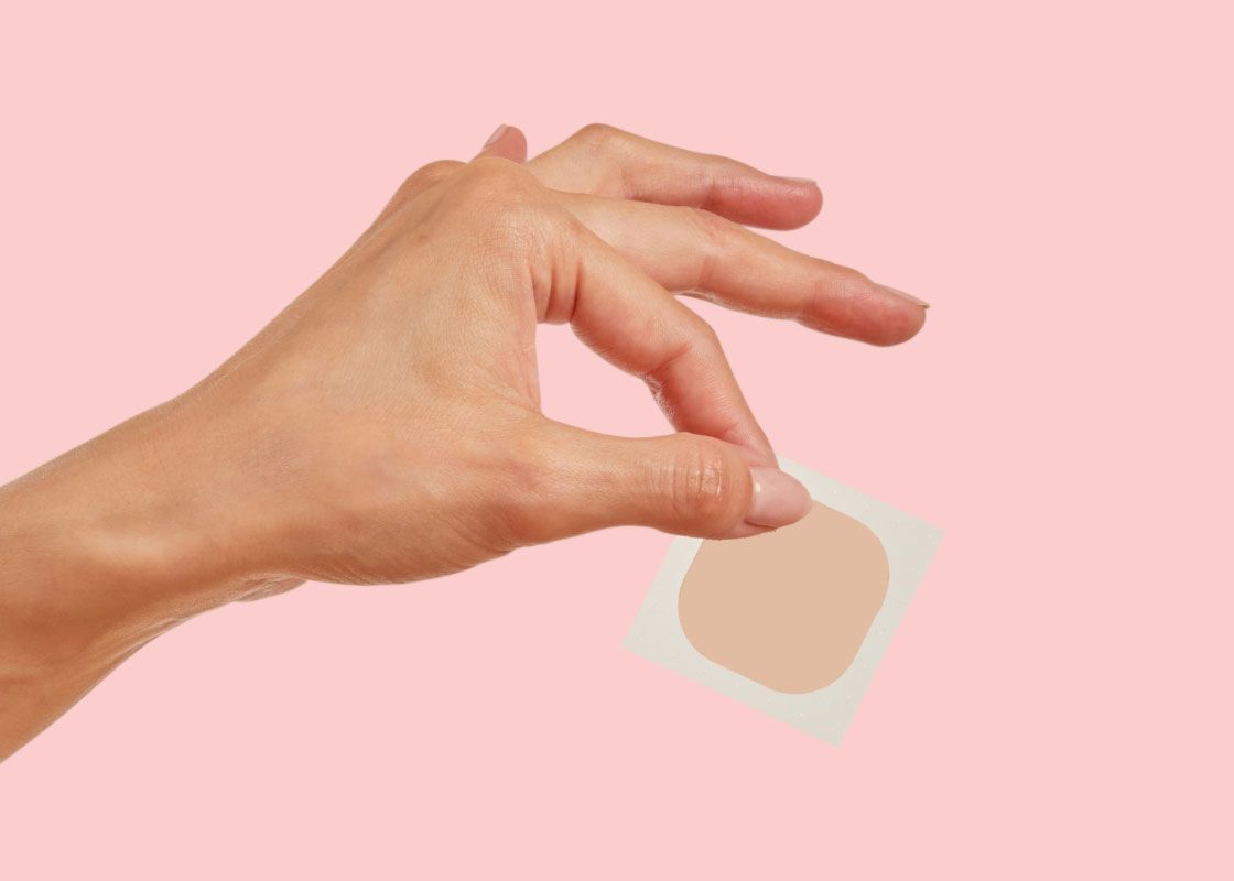 A woman's hand holding an Estradiol patch in front of a pink background