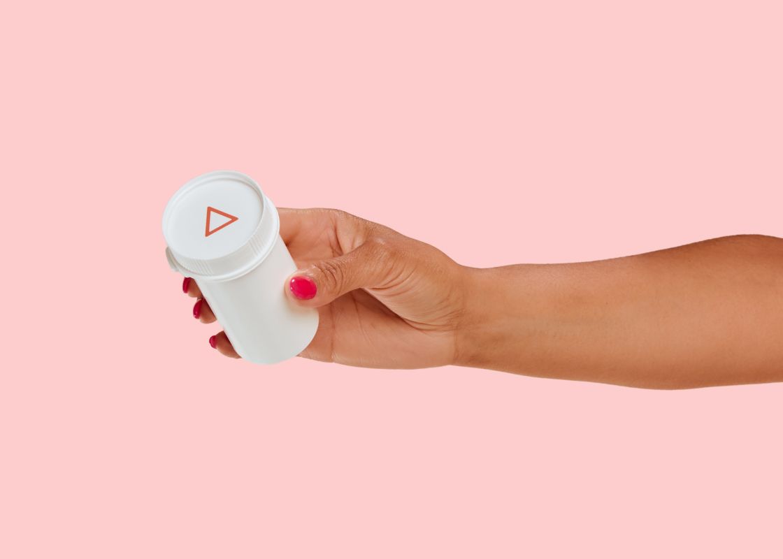 A woman's hand holding a small Wisp pill vial in front of a pink background