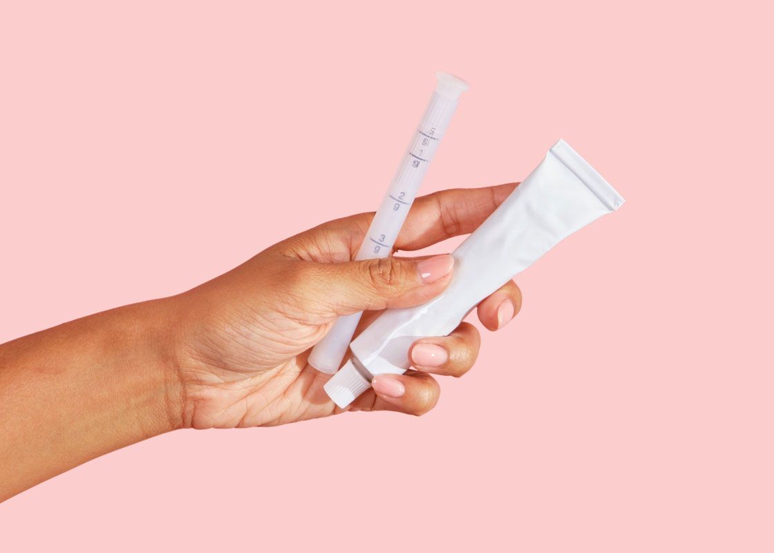 A woman's hand holding a tube of vaginal dryness cream and an applicator in front of a pink background