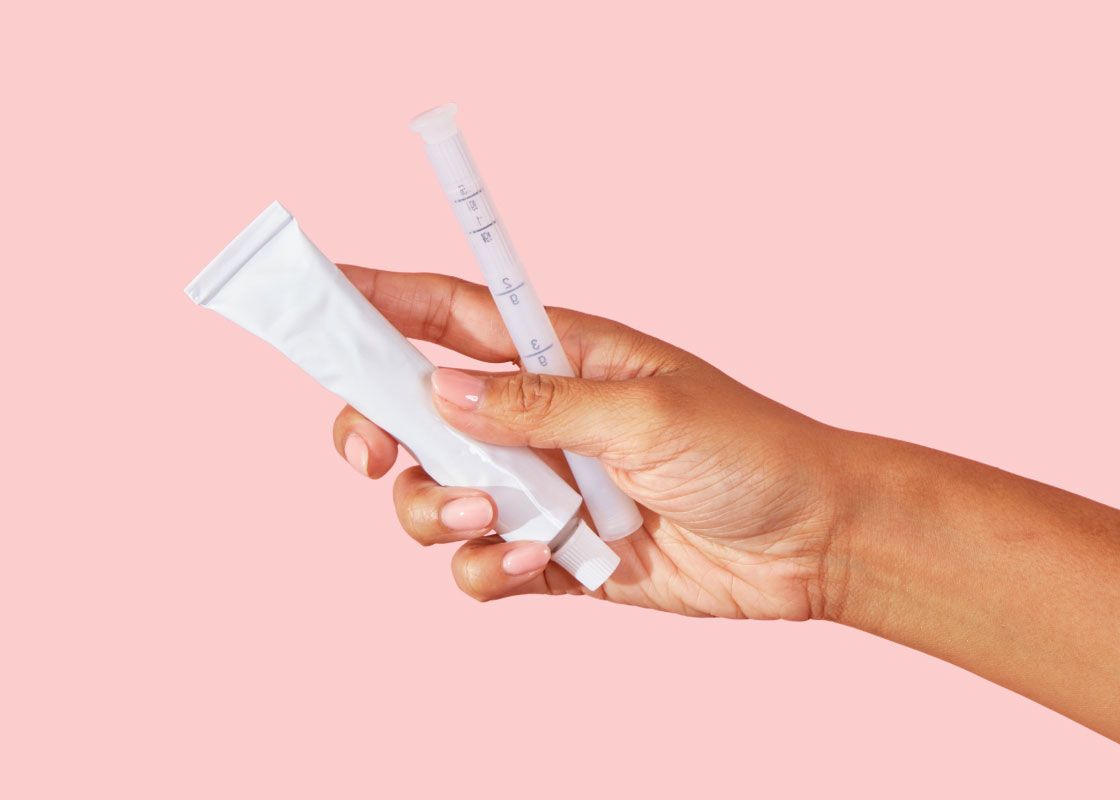 A woman's hand holding a tube of vaginal dryness cream and an applicator in front of a pink background