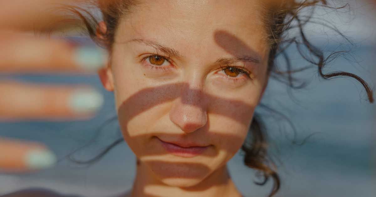 Close-up of a woman's eyes and forehead, with wet hair spread around her head, lit by warm sunlight casting soft shadows across her face.