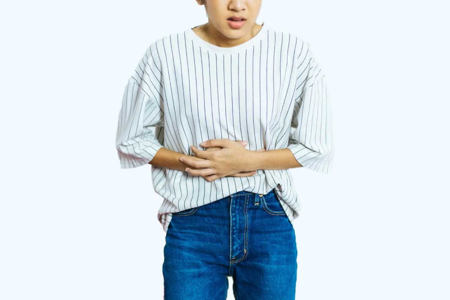 A woman wearing a striped white and black shirt with blue jeans has her hands over her abdomen in obvious discomfort