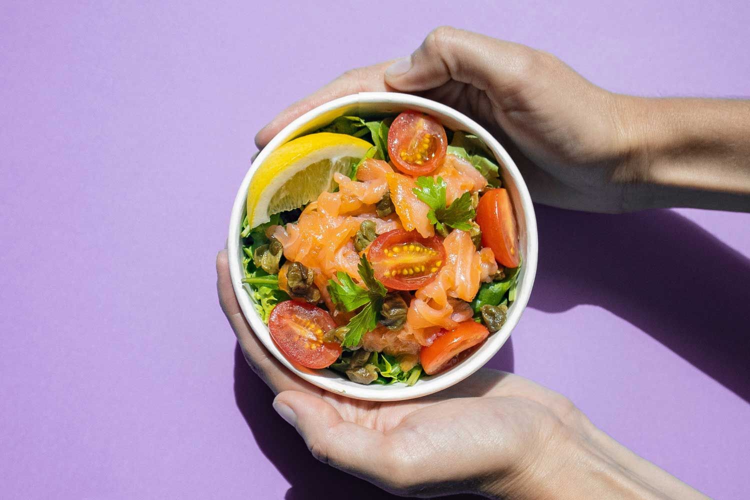 A person's hands holding a bowl of fruits and veggies on a purple surface
