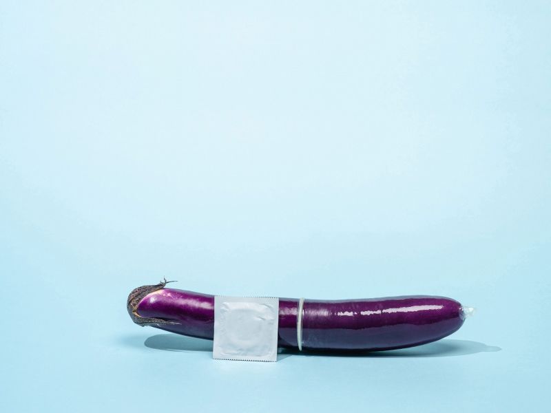 An eggplant with a condom on it and a silver unopened condom wrapper on a light blue surface