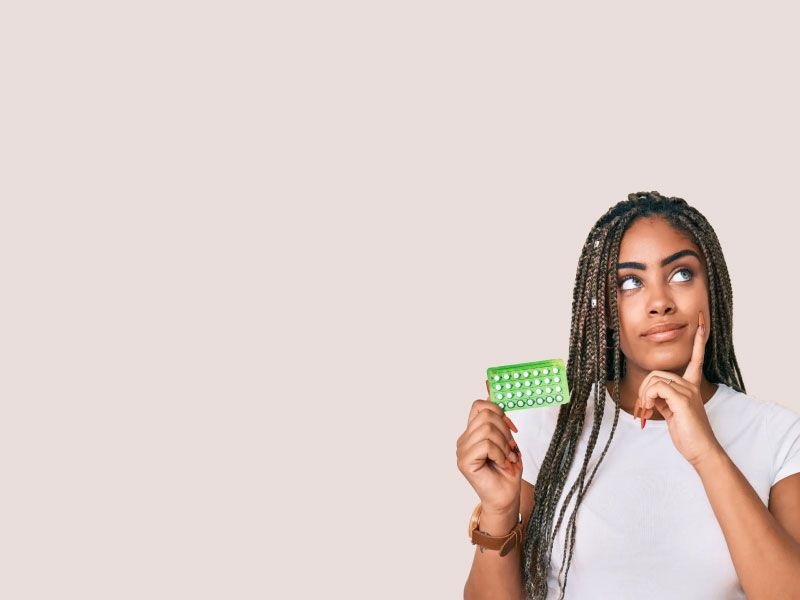 A woman wearing a white shirt and holding a green birth control packet with an expressing of contemplation on her face