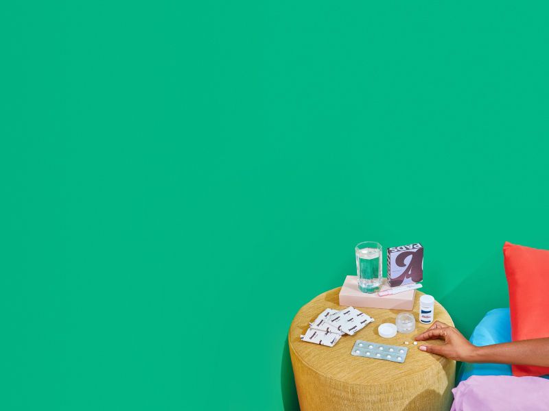 A woman's hand reaching for pills on a yellow nightstand with Wisp's medication abortion kit in front of a green wall with red, blue and pink bedding