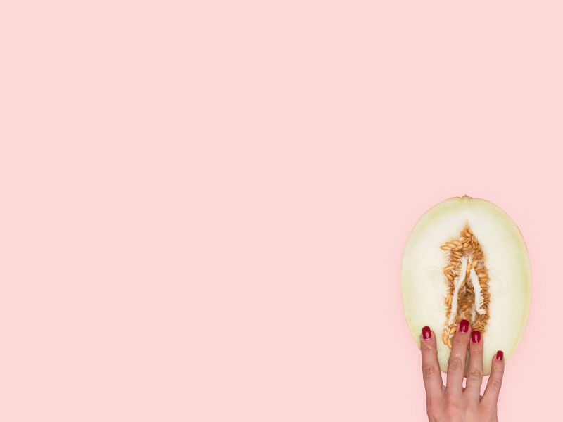 A woman's hand touching a vulva-shaped cut open fruit on a pink surface