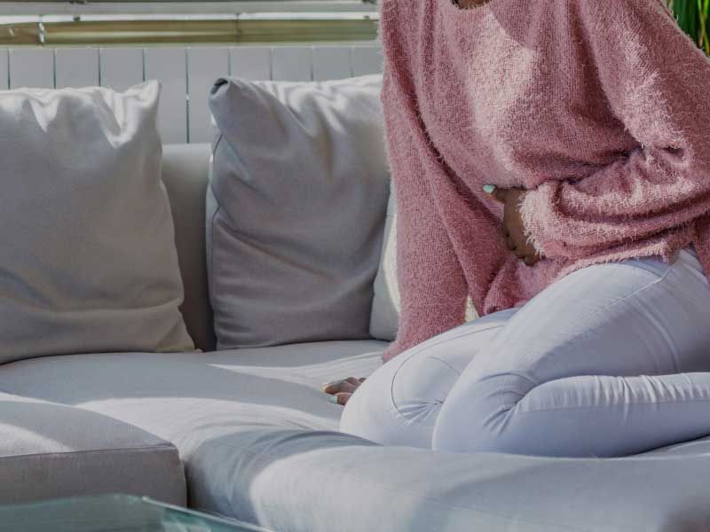 A woman wearing a swearing a pink sweater with her hand on her abdomen in discomfort while sitting on a white couch