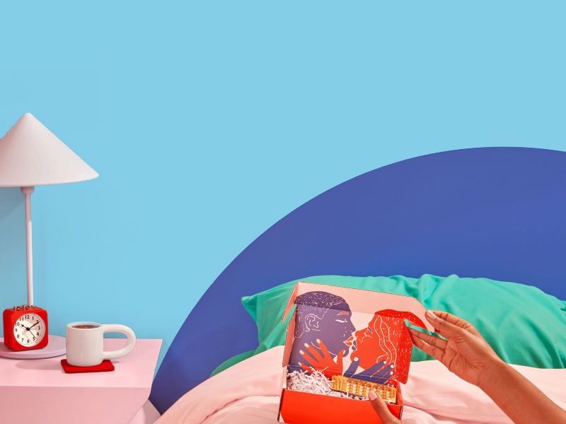 A woman laying on a bed holding a Wisp box with 3 birth control packets in it next to a pink nightstand with a coffee mug, a red alarm clock and a white lamp