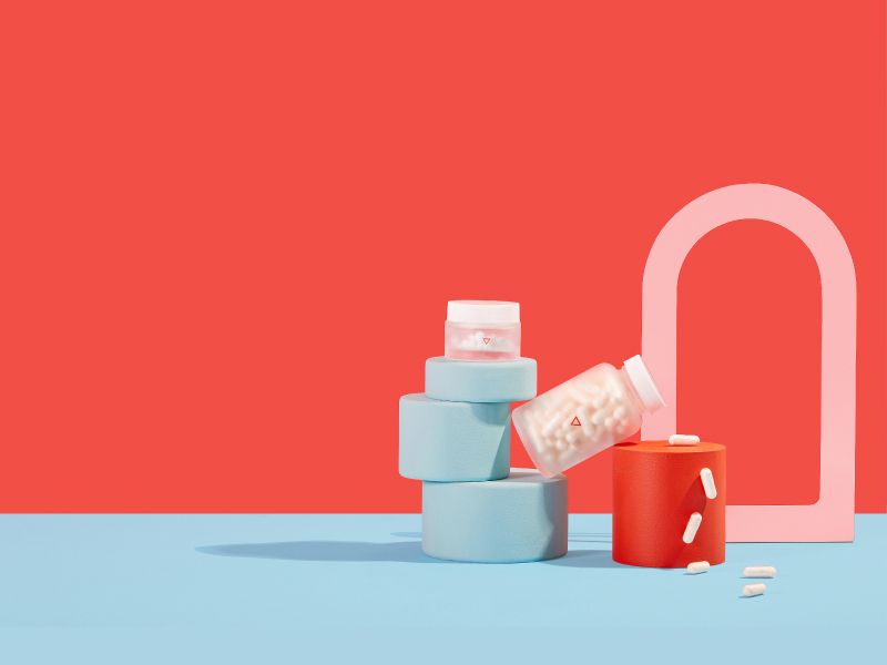 A tall Wisp glass jar with pills pouring out balanced on colorful abstract shapes with a light blue and red background