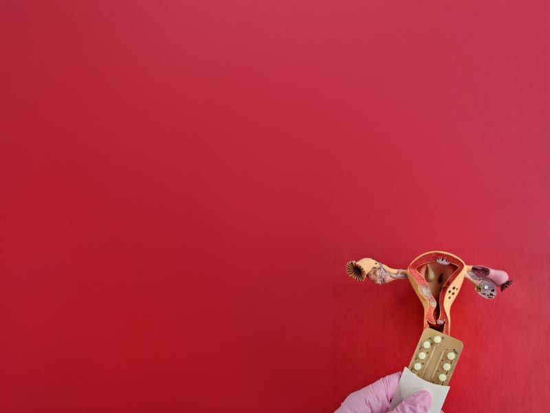 A reproductive organ diagram with a a person's hand holding a birth control packet on a dark red surface