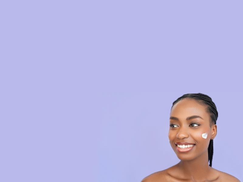 A pretty woman is smiling with skincare cream swiped on her cheeks in front of a purple background