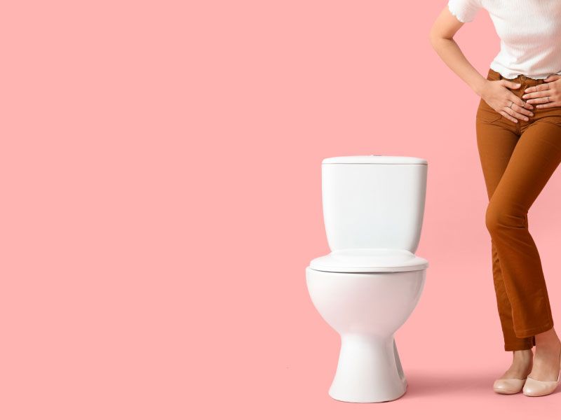 A woman standing next to a toilet wearing a white shirt and brown pants with her hands on her pelvic region indicating discomfort