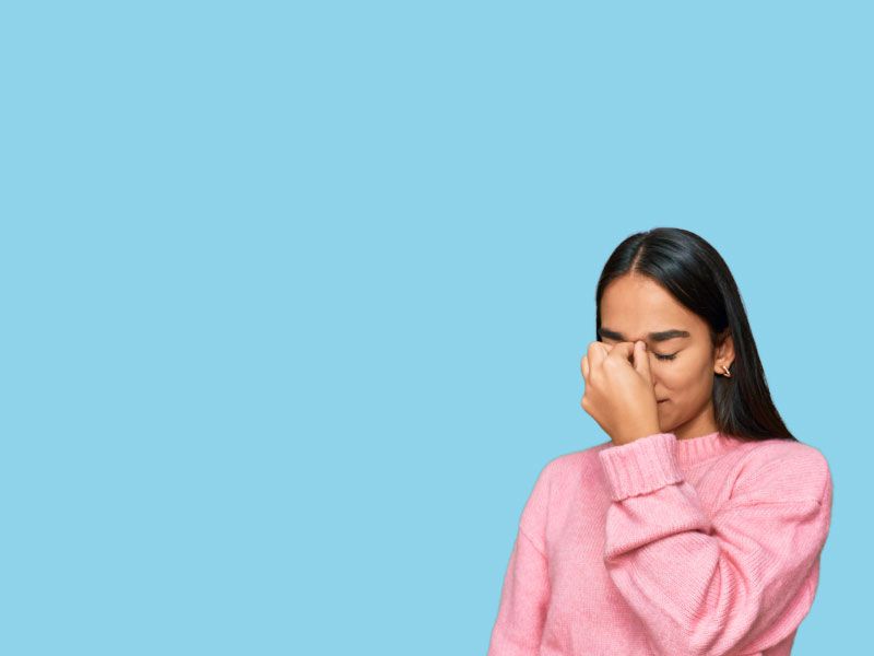 A woman with dark hair and a pink sweater is pinching the bridge of her nose in frustration