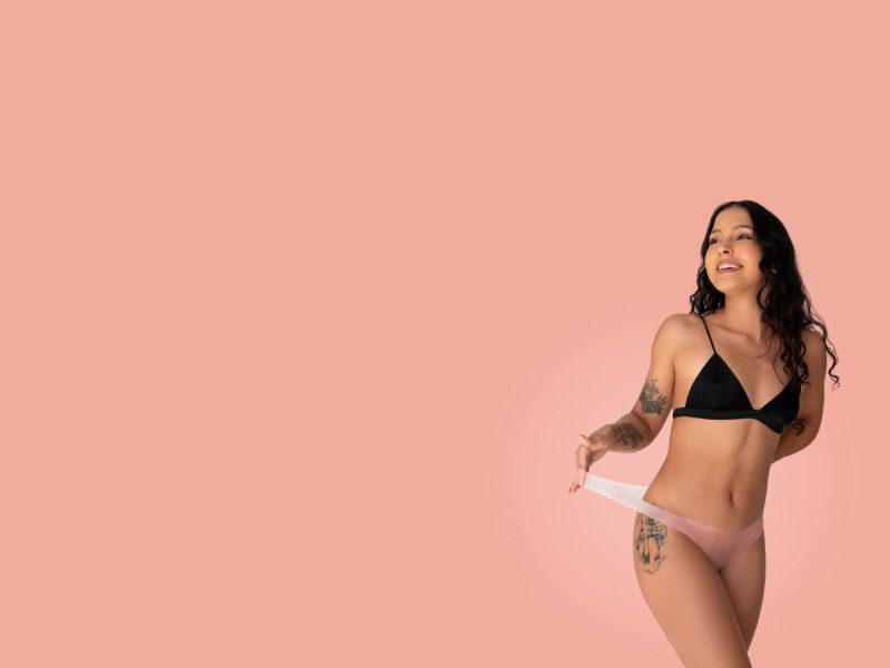 A female wearing a black bra and latex Lorals underwear in front of a peach background