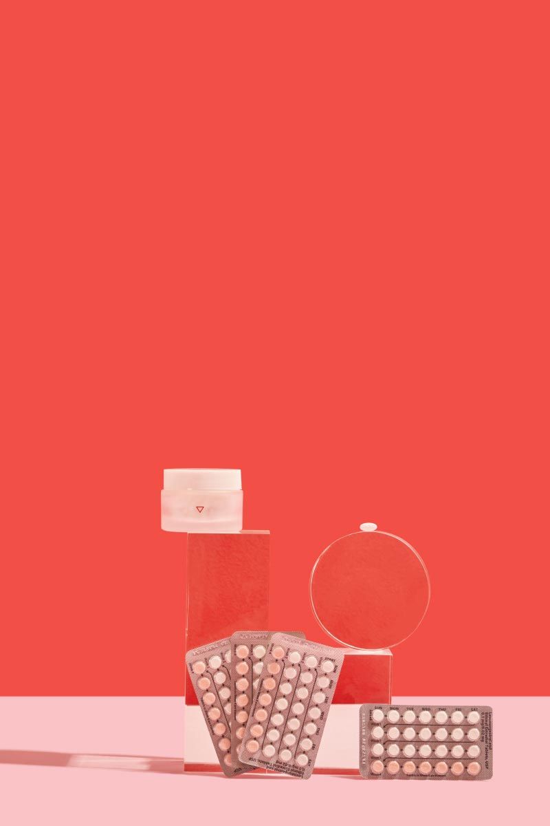 4 birth control packets and a small glass pill jar balanced on clear abstract shapes on a pink surface with a red background