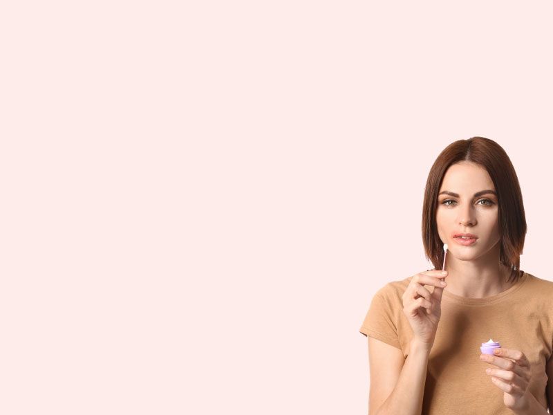 A woman with brown hear and a tan shirt is applying cream to a cold sore with a cotton swab