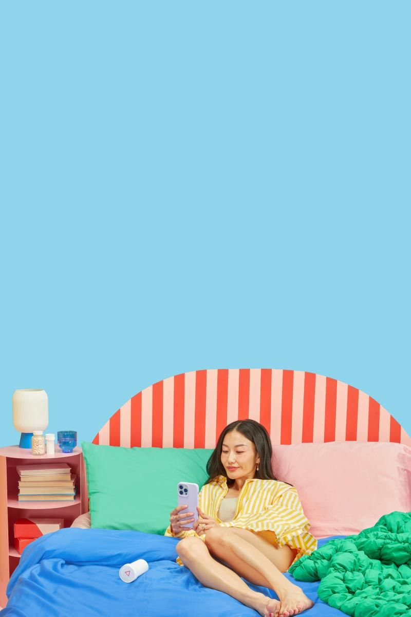 A woman laying on a bed with colorful bedding using a mobile phone next to a nightstand with Wisp medication bottles